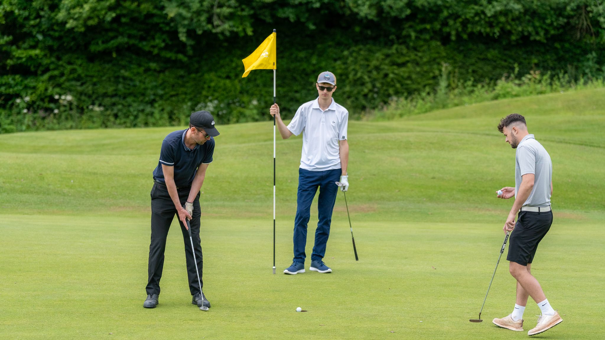 Three men playing golf on a green, one putting, one holding the flag, and another preparing his putt.