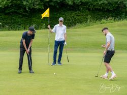 Three men playing golf on a green, one putting, one holding the flag, and another preparing his putt.
