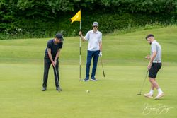 Three men playing golf on a green, one putting, one holding the flag, and another preparing his putt.