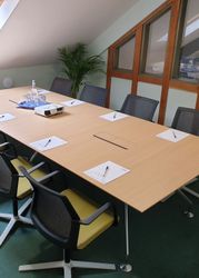 Modern meeting room with a long wooden table, eight chairs, notepads with pens, a projector, and glassware under a skylight.