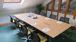 Modern meeting room with a long wooden table, eight chairs, notepads with pens, a projector, and glassware under a skylight.