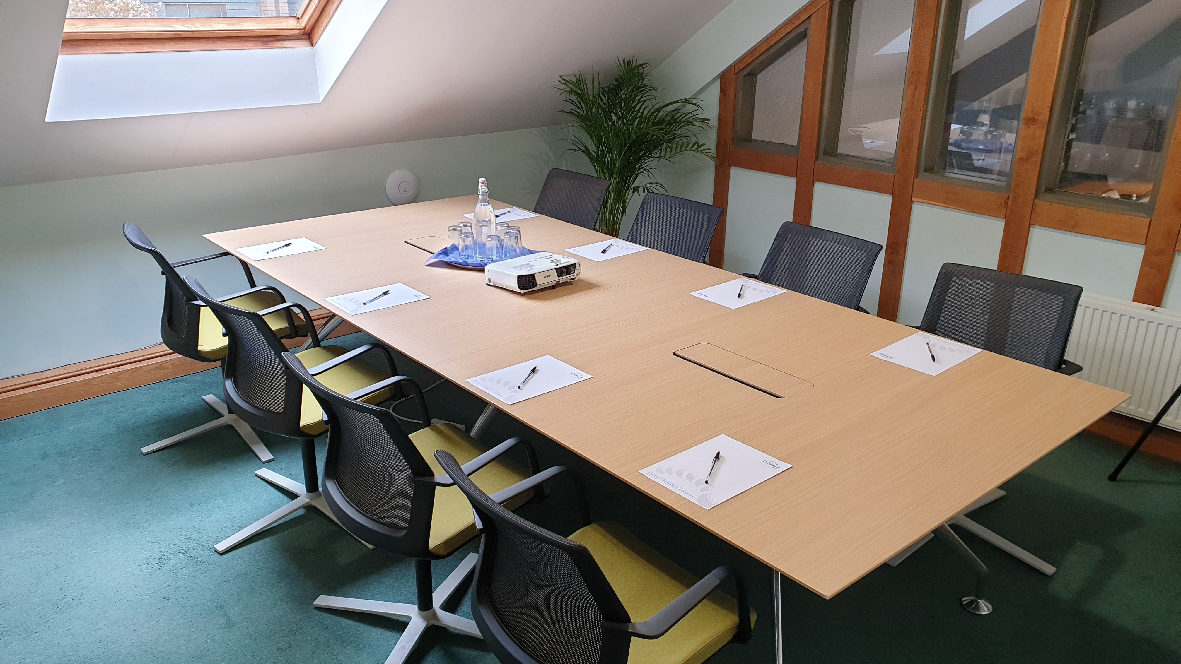Modern meeting room with a long wooden table, eight chairs, notepads with pens, a projector, and glassware under a skylight.