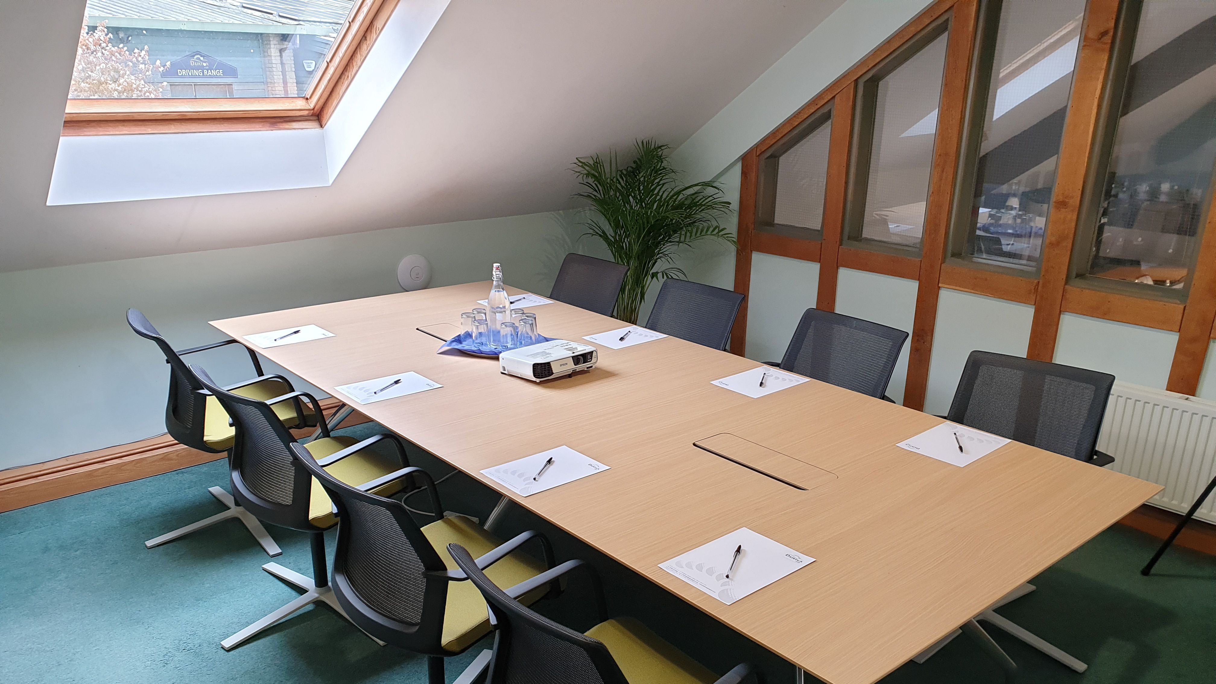 Modern meeting room with a long wooden table, eight chairs, notepads with pens, a projector, and glassware under a skylight.