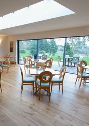 Wide angle view of a modern orangery with wooden tables and chairs, large windows, and garden scenery outside