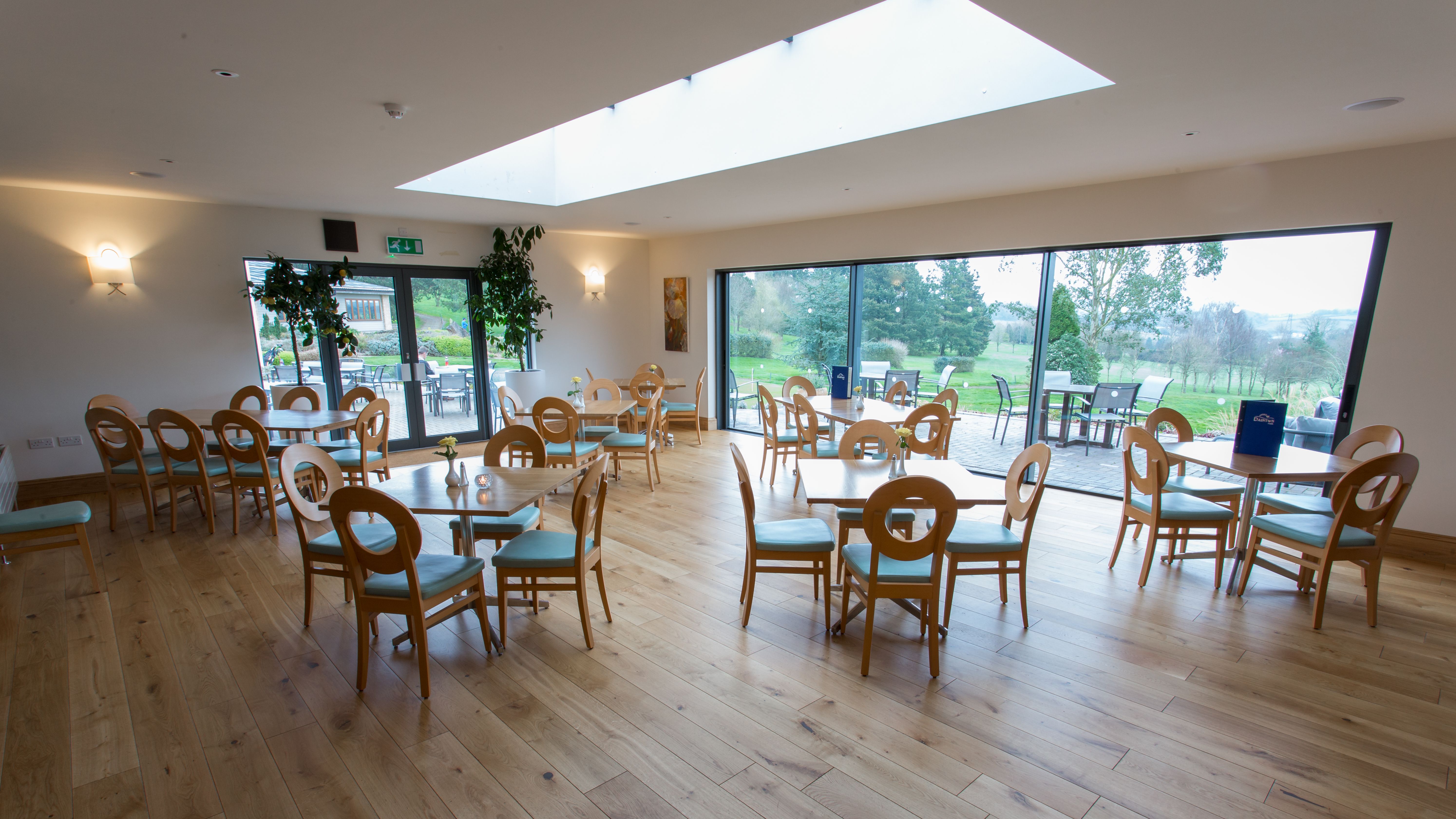 Wide angle view of a modern orangery with wooden tables and chairs, large windows, and garden scenery outside
