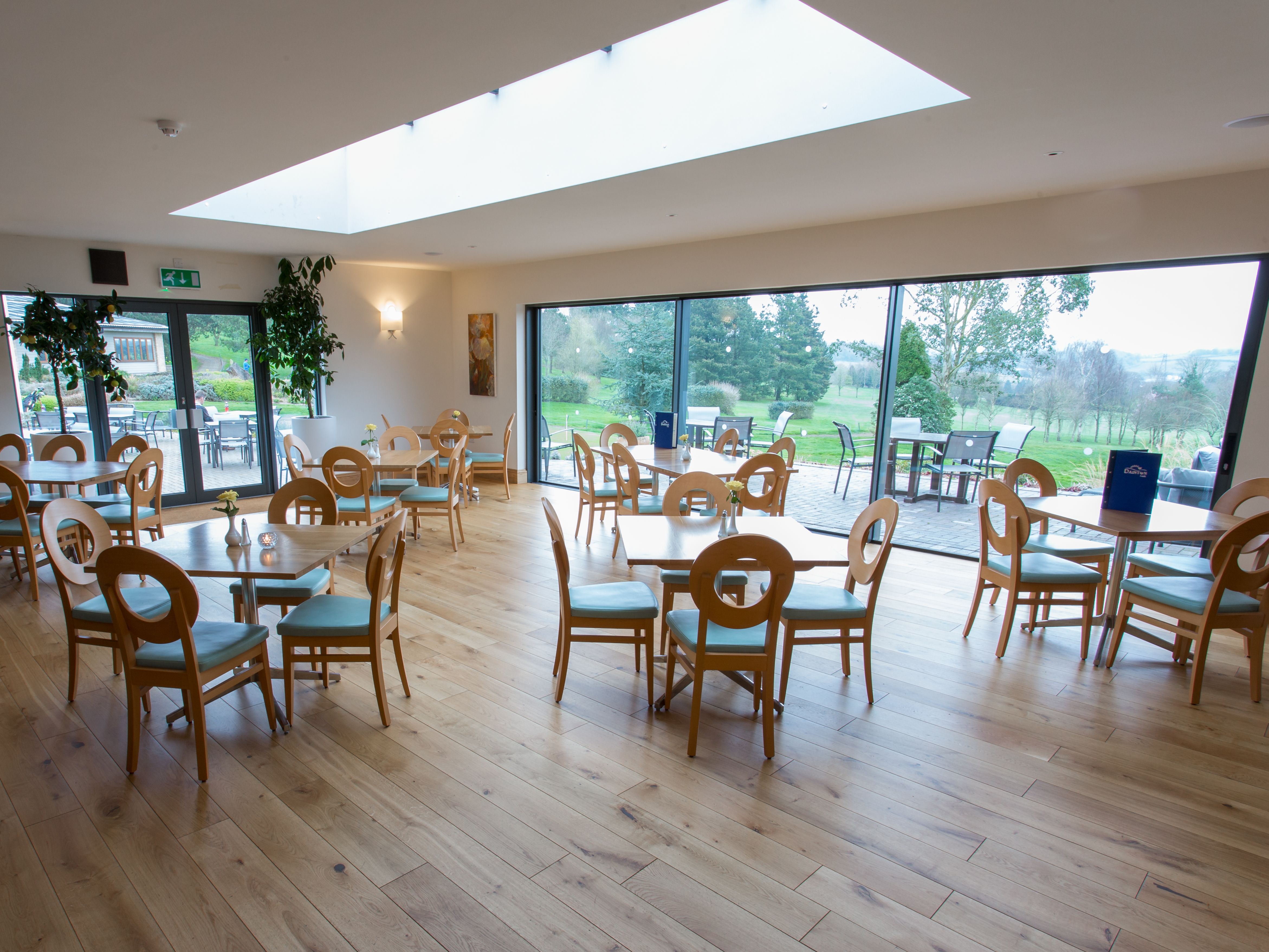 Wide angle view of a modern orangery with wooden tables and chairs, large windows, and garden scenery outside