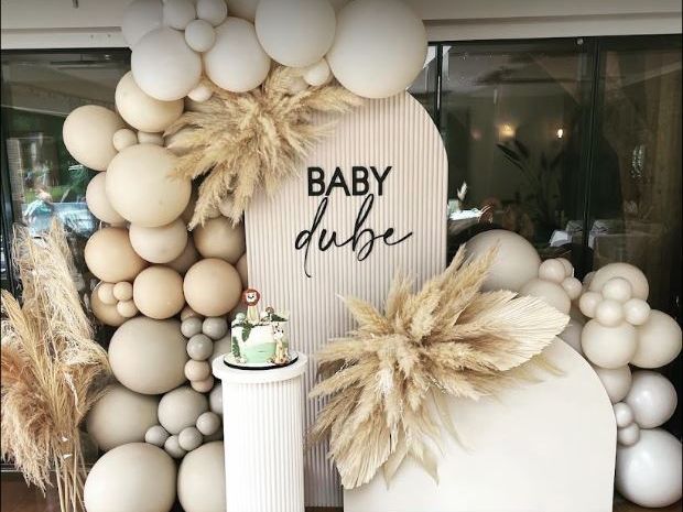 A neutral-toned baby shower setup with balloon decorations, pampas grass, and a cake on a white pedestal. The backdrop says 'Baby dube'.