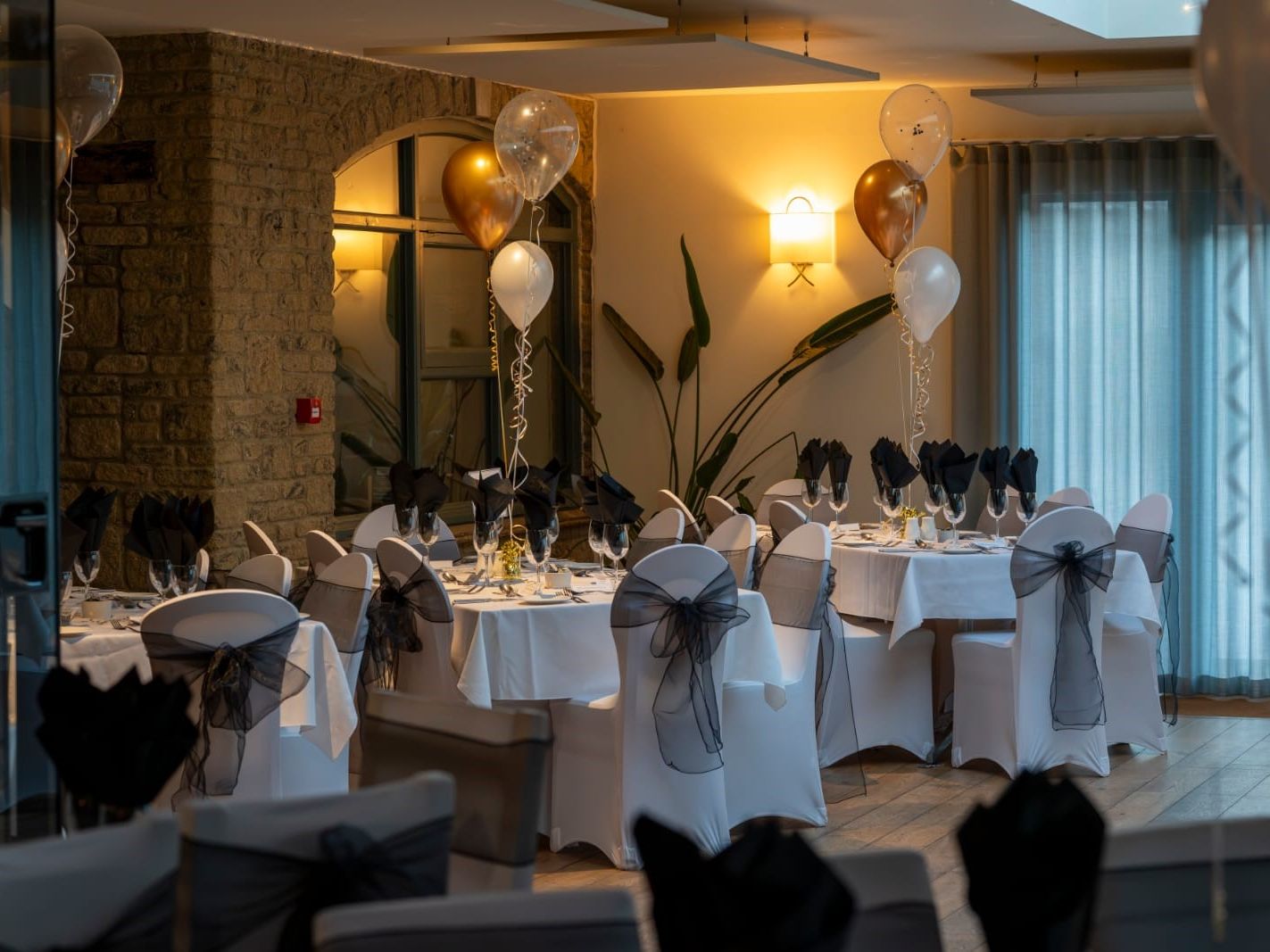 Elegant indoor banquet setup with round tables, white tablecloths, black napkins, and gold and white balloons.