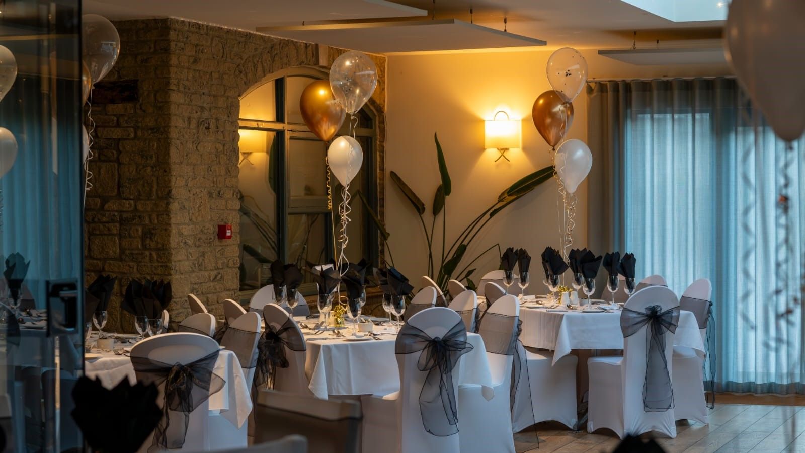 Elegant indoor banquet setup with round tables, white tablecloths, black napkins, and gold and white balloons.