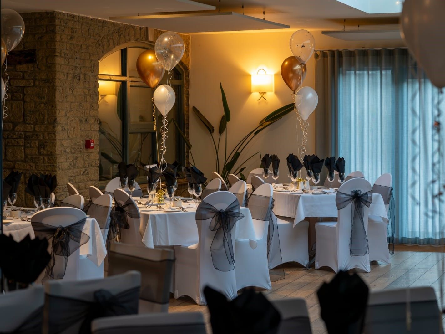 Elegant indoor banquet setup with round tables, white tablecloths, black napkins, and gold and white balloons.
