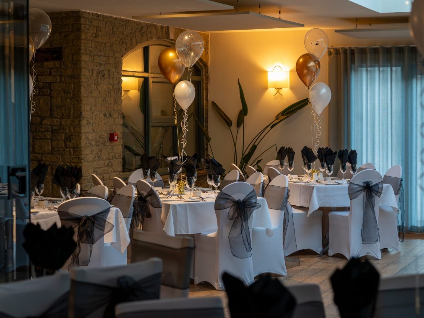 Elegant indoor banquet setup with round tables, white tablecloths, black napkins, and gold and white balloons.