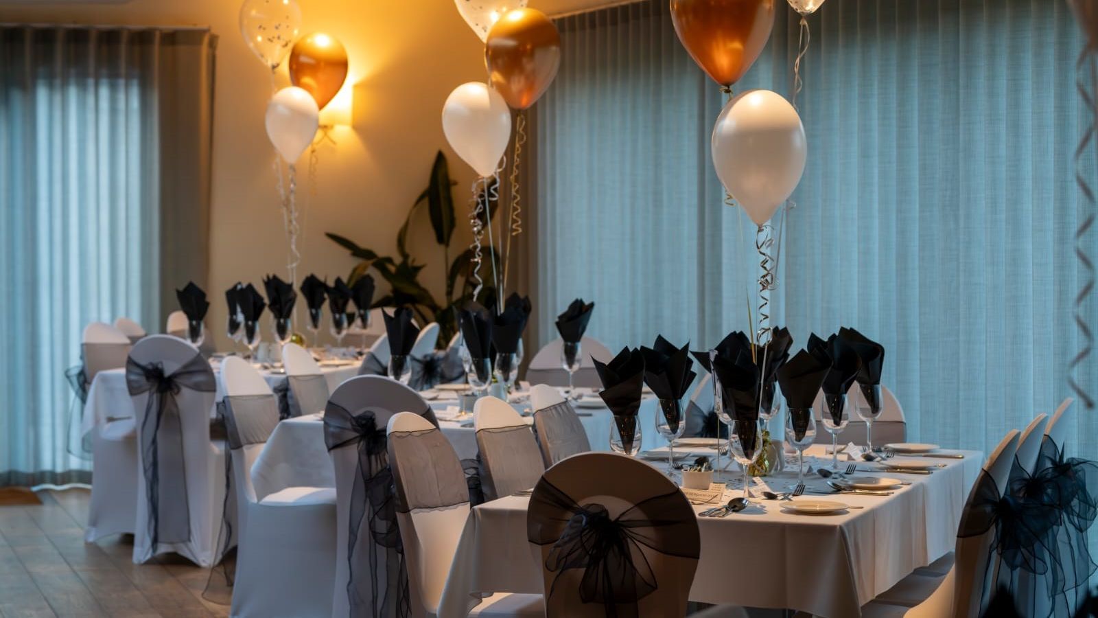 Elegant banquet room with white tablecloths, black napkins, and gold, white, and clear balloons as decorations.