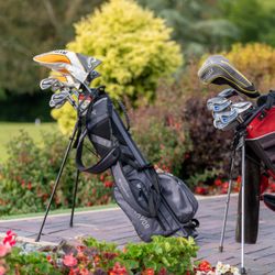 Two golf bags with clubs standing on a paved path near a golf course surrounded by greenery and flowers.