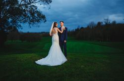 Bride and groom standing on a green lawn at dusk with trees and a dramatic blue sky in the background.