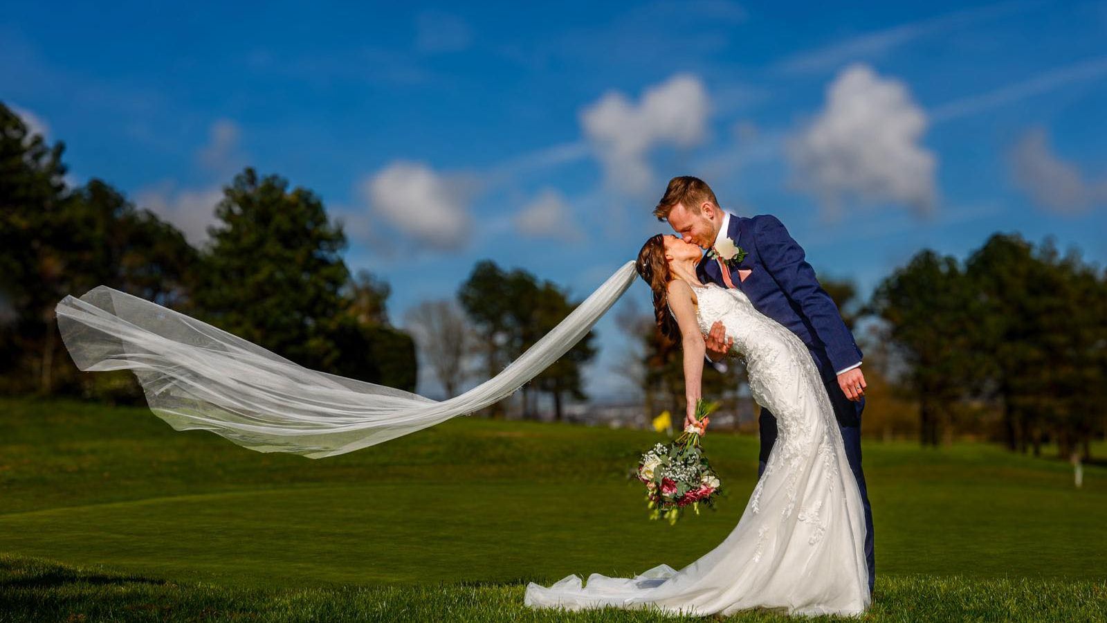 Bride and groom sharing a romantic kiss outdoors, with the bride's veil flowing in the wind.