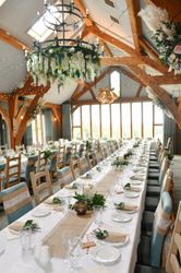 Long banquet tables set for a wedding reception inside a rustic venue with wooden beams and floral decorations.