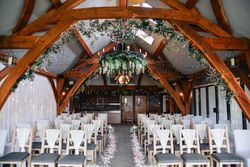 Beautifully decorated indoor wedding ceremony setup with rows of chairs, floral aisle, fairy lights, and greenery hanging from wooden beams.