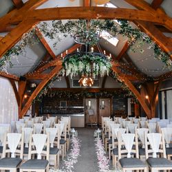 Beautifully decorated indoor wedding ceremony setup with rows of chairs, floral aisle, fairy lights, and greenery hanging from wooden beams.