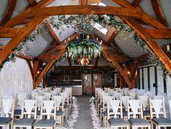 Beautifully decorated indoor wedding ceremony setup with rows of chairs, floral aisle, fairy lights, and greenery hanging from wooden beams.