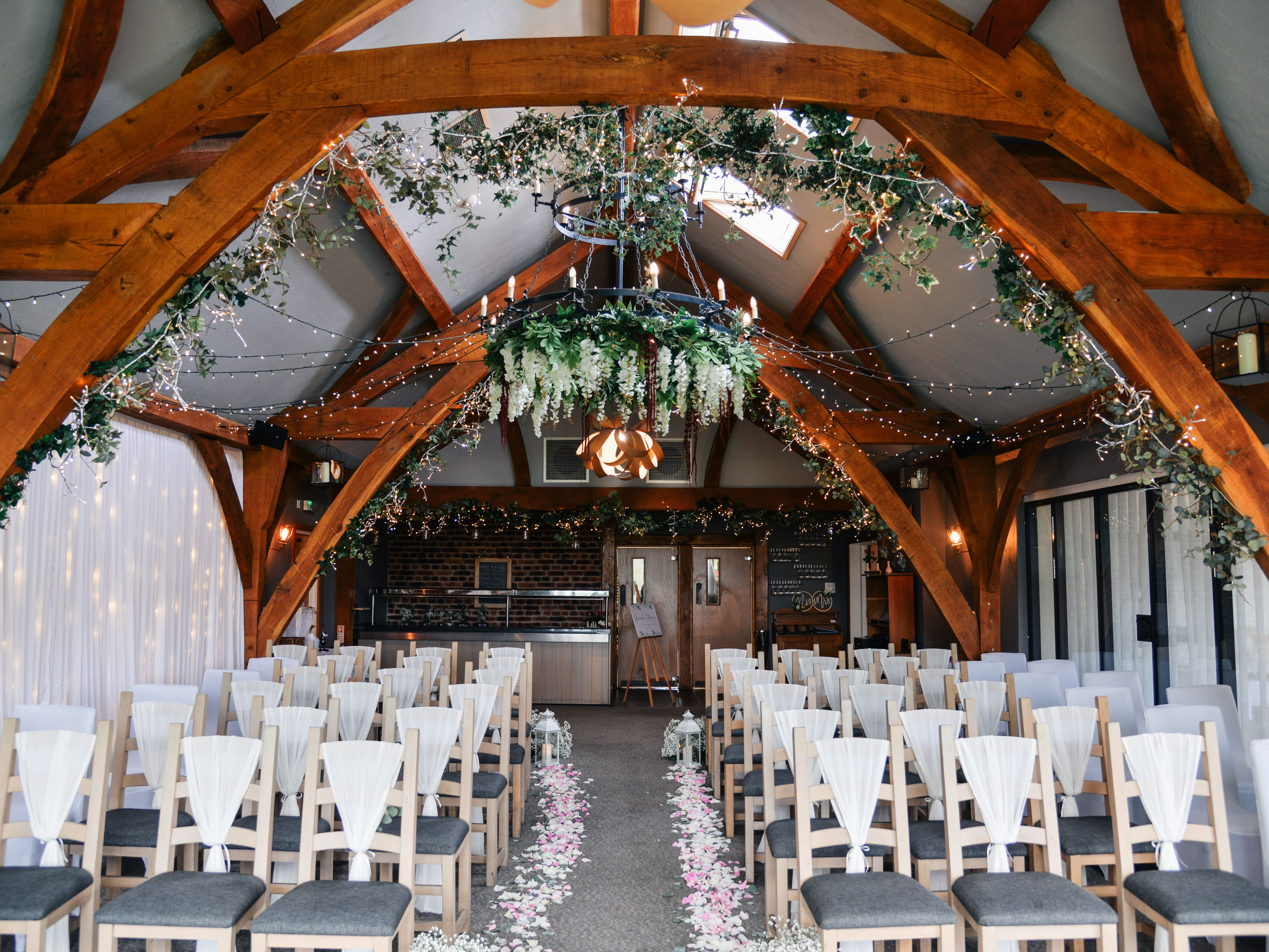 Beautifully decorated indoor wedding ceremony setup with rows of chairs, floral aisle, fairy lights, and greenery hanging from wooden beams.