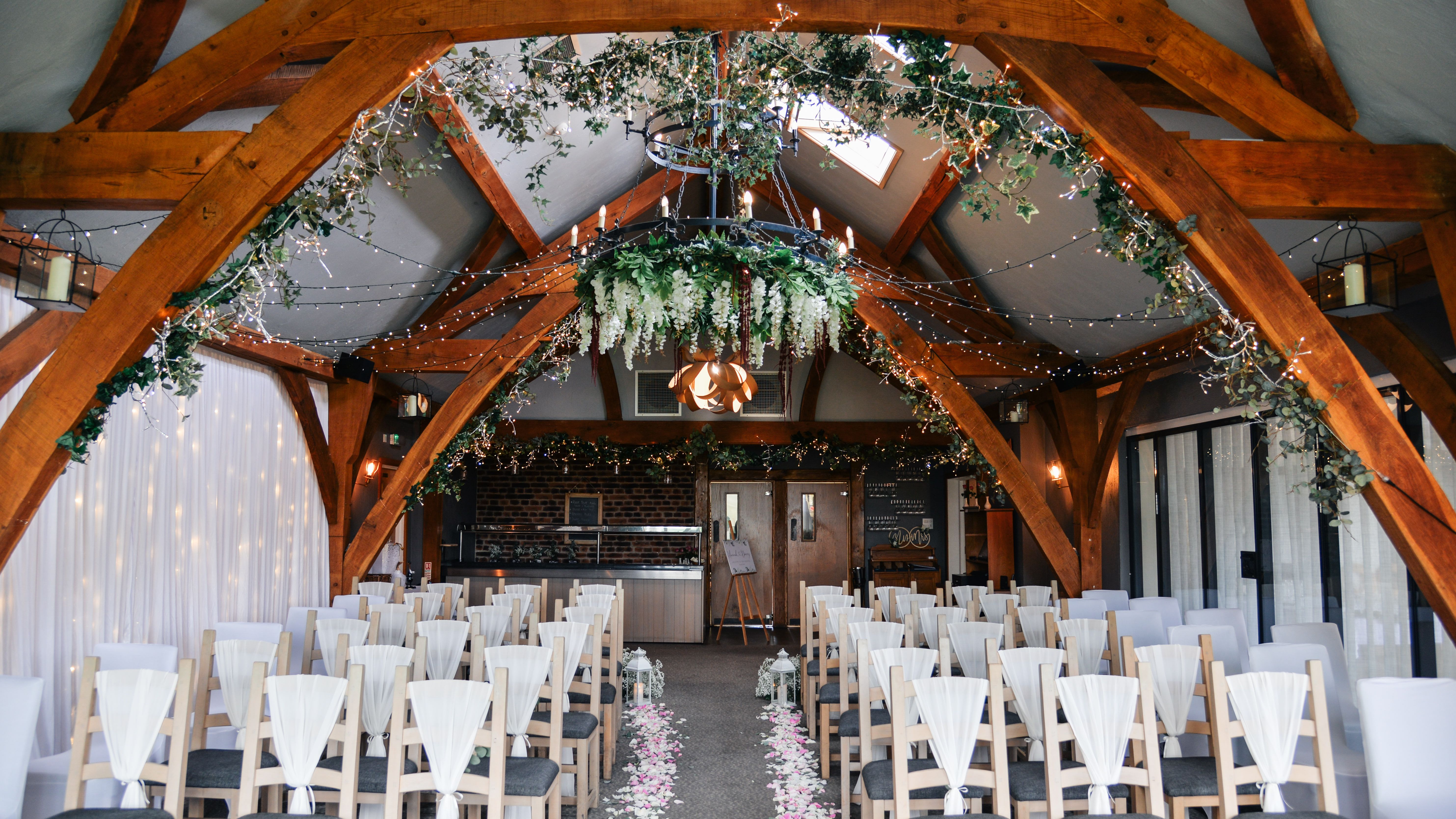 Beautifully decorated indoor wedding ceremony setup with rows of chairs, floral aisle, fairy lights, and greenery hanging from wooden beams.