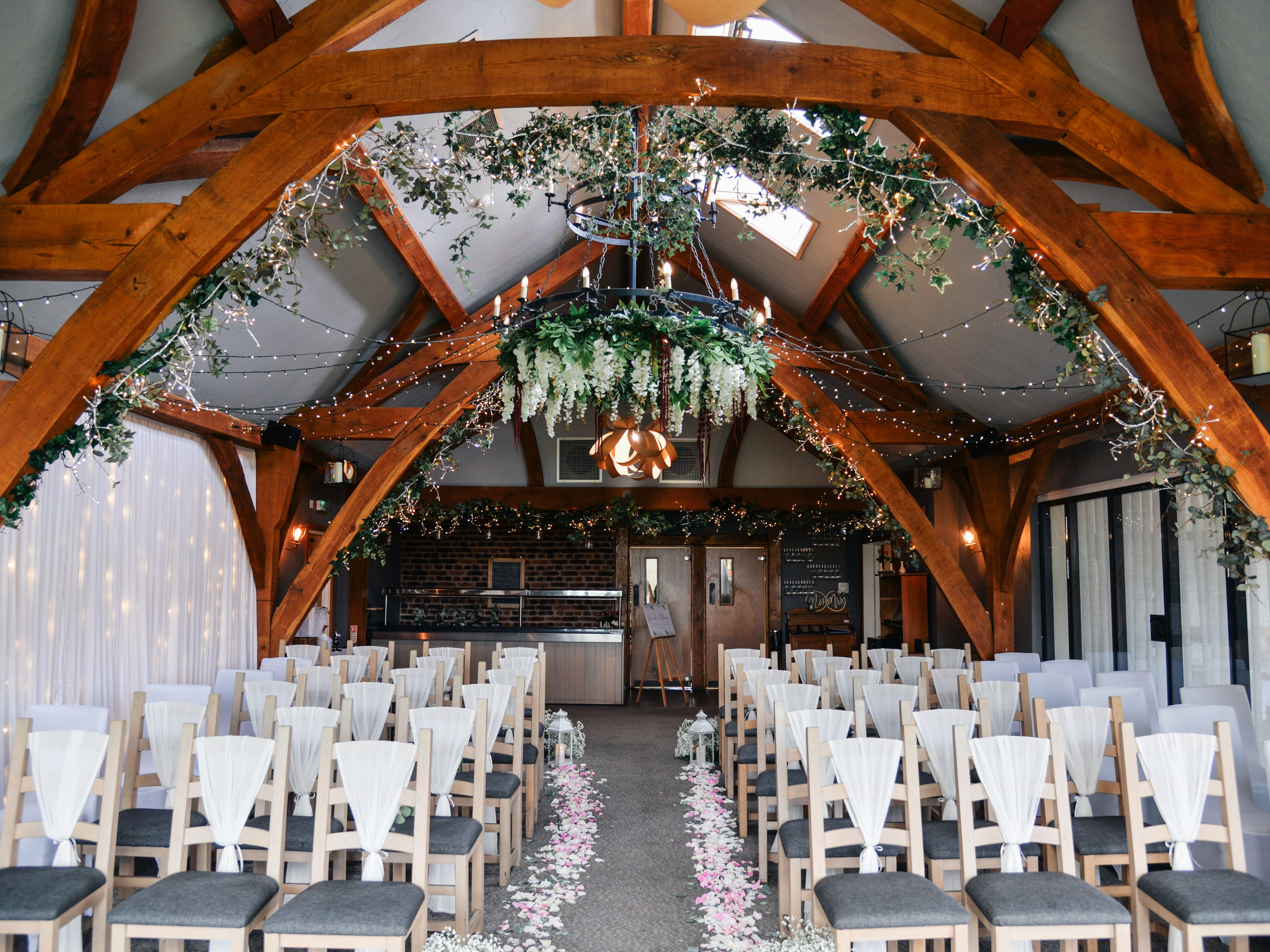 Beautifully decorated indoor wedding ceremony setup with rows of chairs, floral aisle, fairy lights, and greenery hanging from wooden beams.