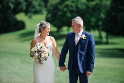 Bride and groom holding hands outdoors, smiling at each other on a sunny day.