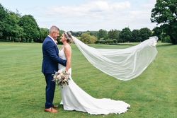 Bride and groom standing on a grassy field, with the bride's veil flowing in the wind.