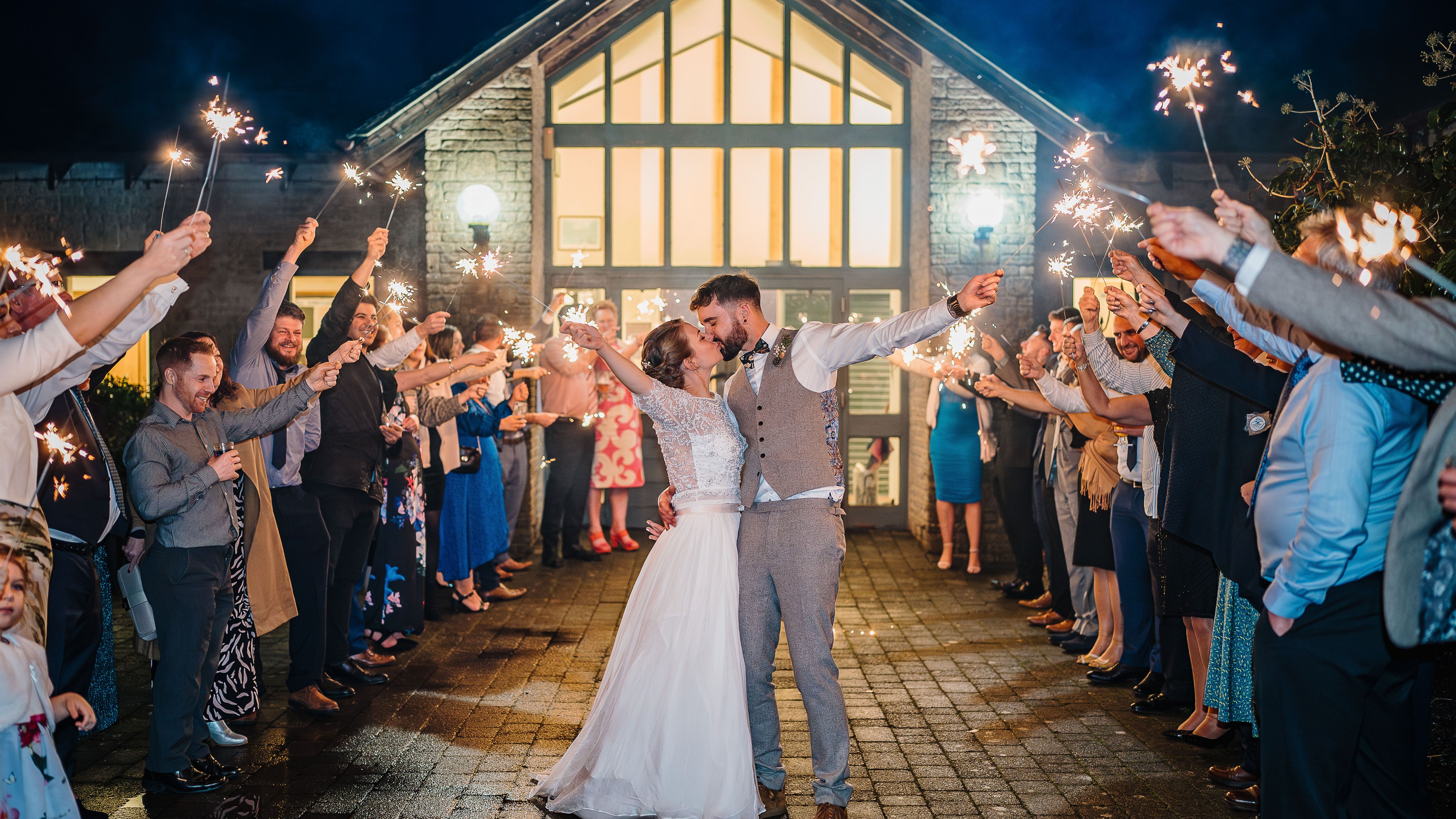 Bride and groom kissing with sparklers held by guests forming an archway outside a venue at night