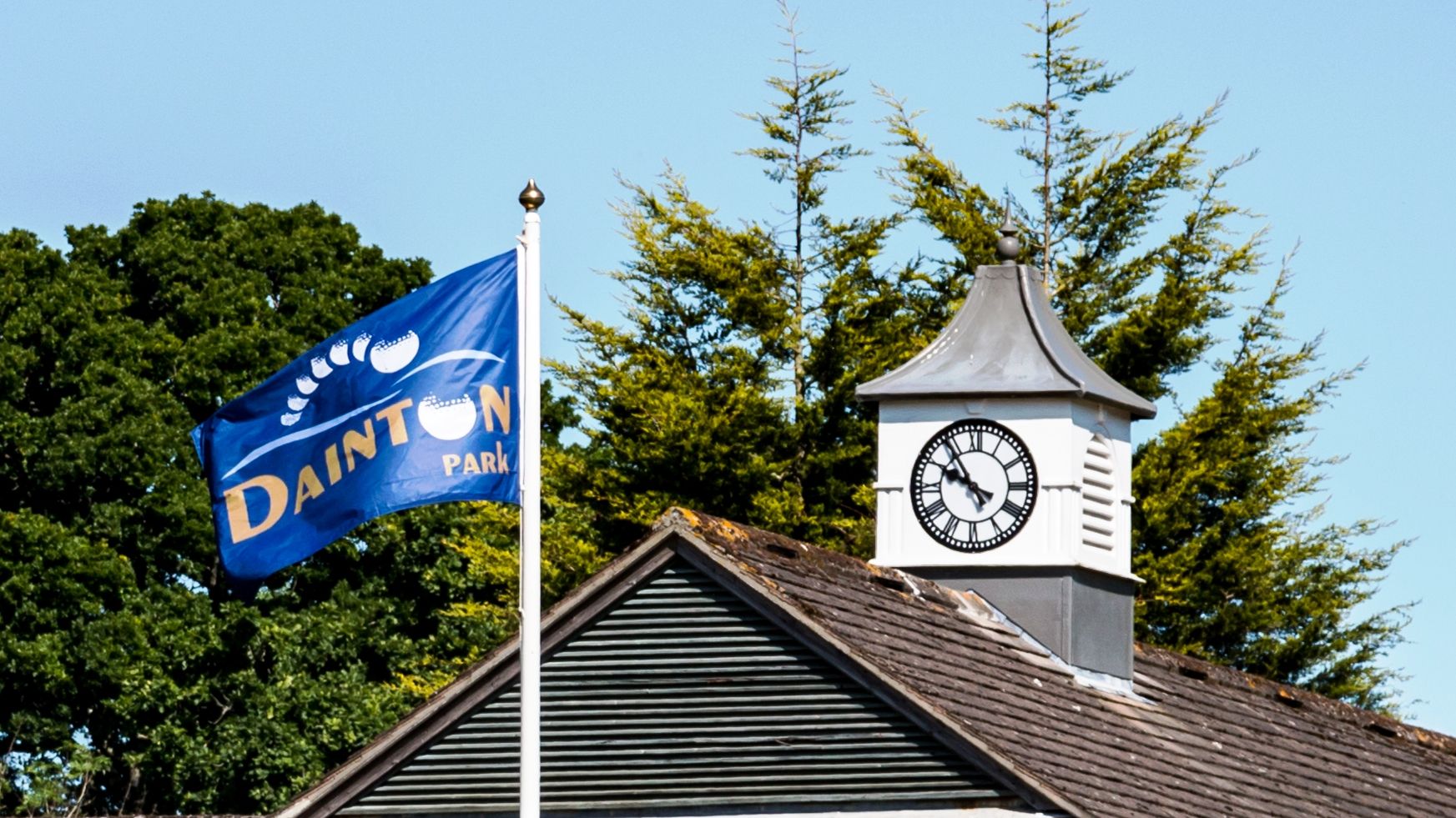 Building with a rooftop clock tower and a flag that reads 'Dainton Park' on a clear day.