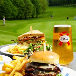 Outdoor table with plates of burgers, sandwiches, chips, and salads with glasses of beer, overlooking a green golf course with people playing in the background.