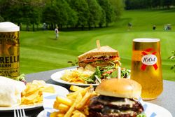 Outdoor table with plates of burgers, sandwiches, chips, and salads with glasses of beer, overlooking a green golf course with people playing in the background.