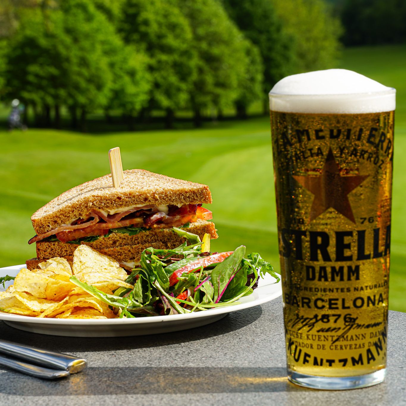 A plate with a sandwich, potato chips, and salad next to a glass of Estrella Damm beer, set on a table outdoors overlooking a green golf course.