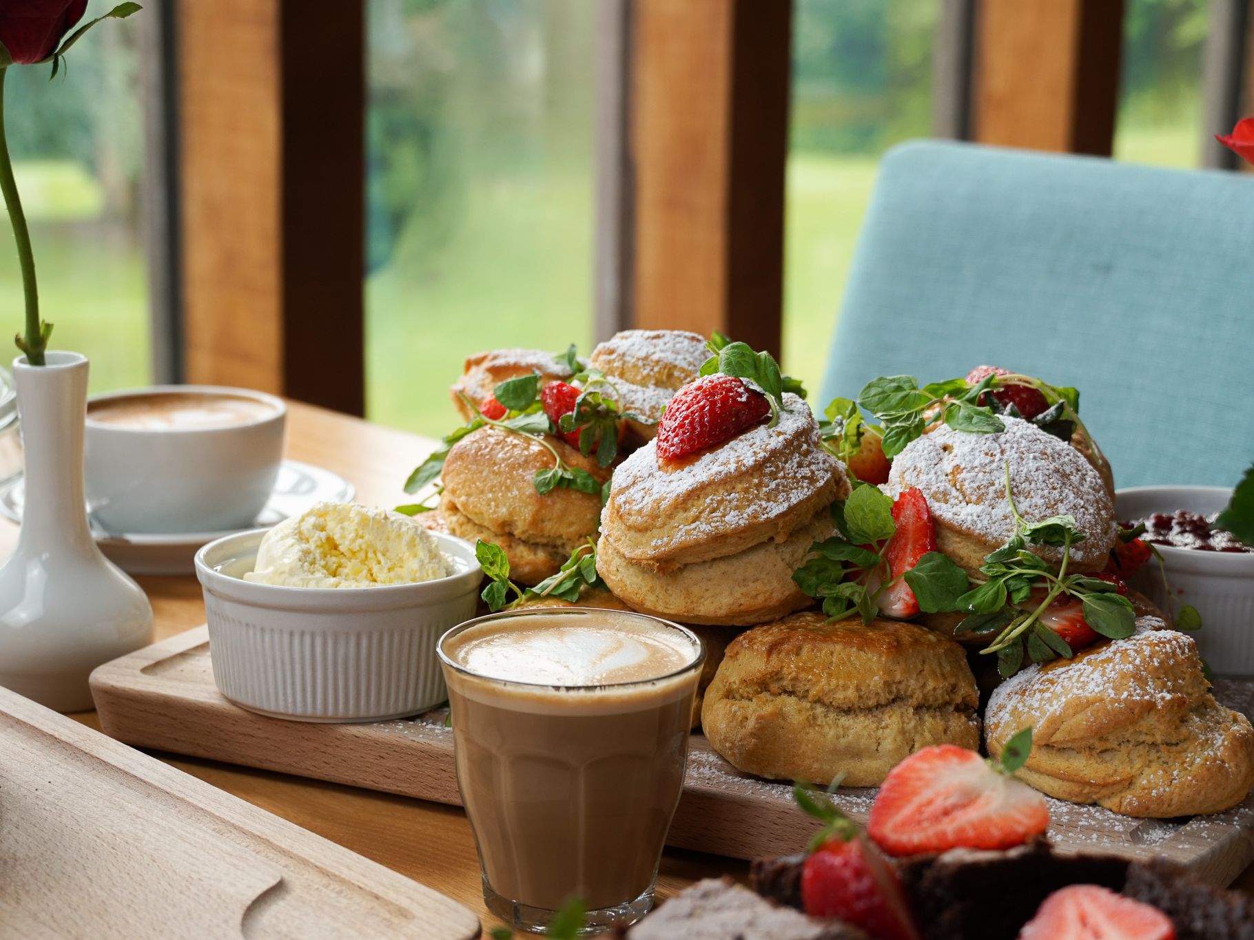 A table set for afternoon tea with scones topped with strawberries and powdered sugar, a glass of coffee, a cup of cappuccino, a bowl of clotted cream, and a vase with a single red rose.