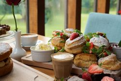 A table set for afternoon tea with scones topped with strawberries and powdered sugar, a glass of coffee, a cup of cappuccino, a bowl of clotted cream, and a vase with a single red rose.