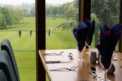 A dining table set with glassware and cutlery by a large window overlooking a golf course where several people are playing golf.