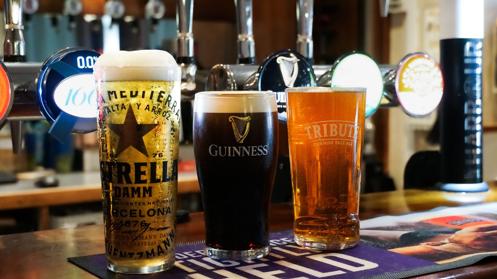 Three pints of beer on a bar, featuring Estrella Damm, Guinness, and Tribute Cornish Pale Ale with beer taps in the background.
