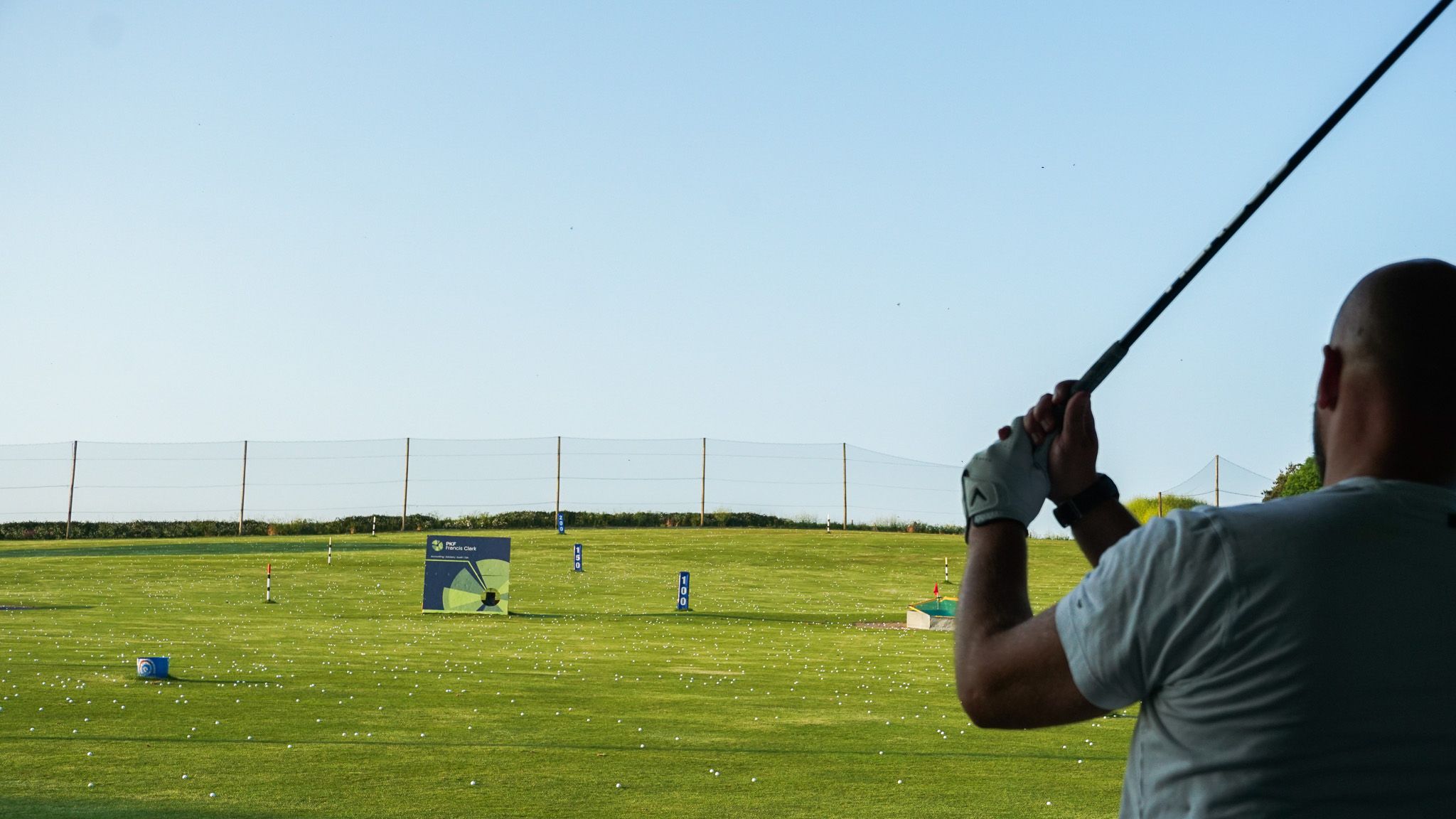 Person swinging a golf club at a driving range with green grass, scattered golf balls, distance markers, and a clear blue sky.