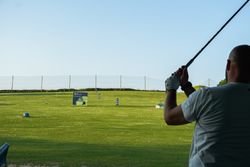 Person swinging a golf club at a driving range with green grass, scattered golf balls, distance markers, and a clear blue sky.