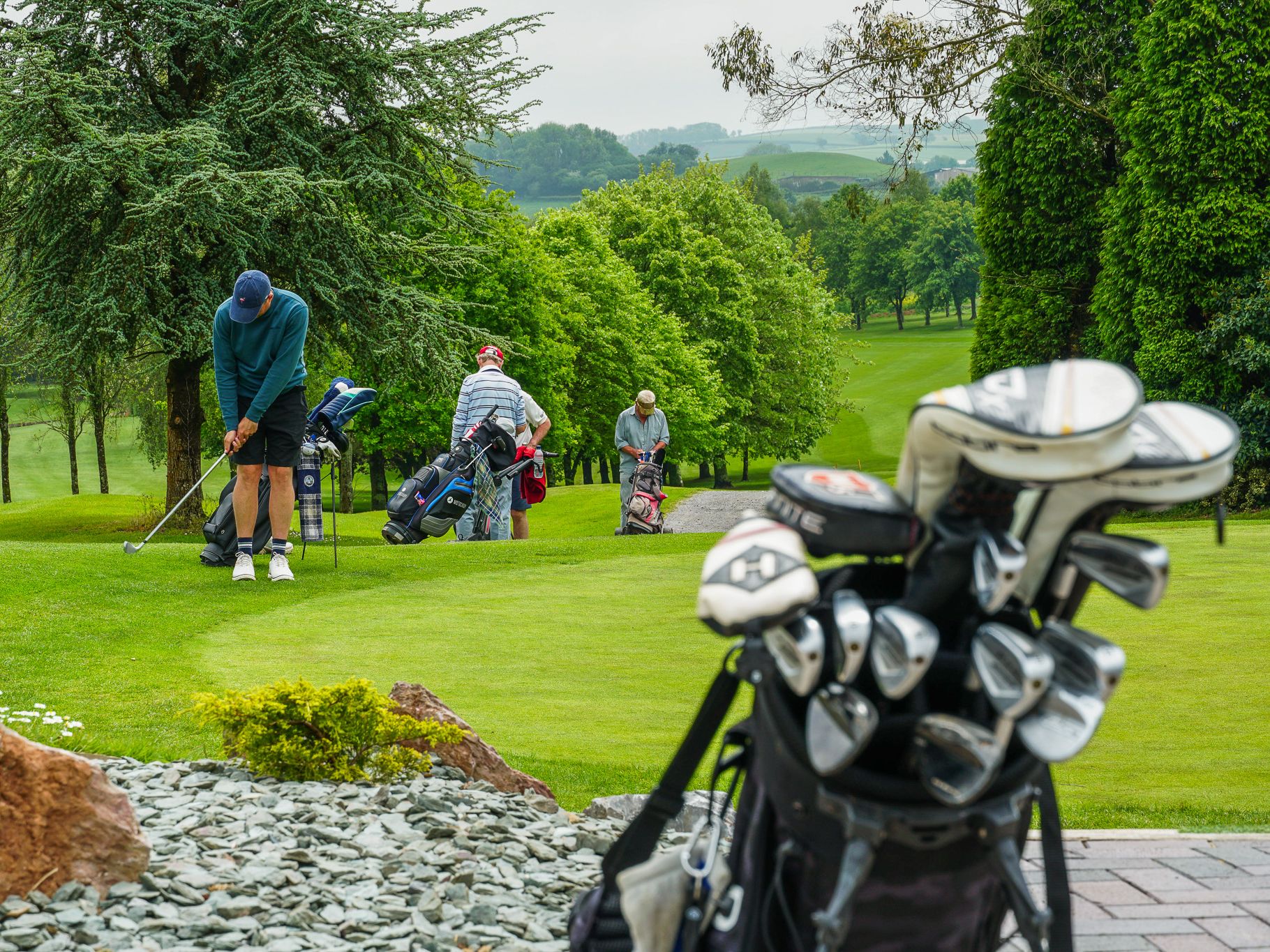 Golfers preparing to play on a green golf course with golf bags and clubs in the foreground. Trees and rolling hills are visible in the background.