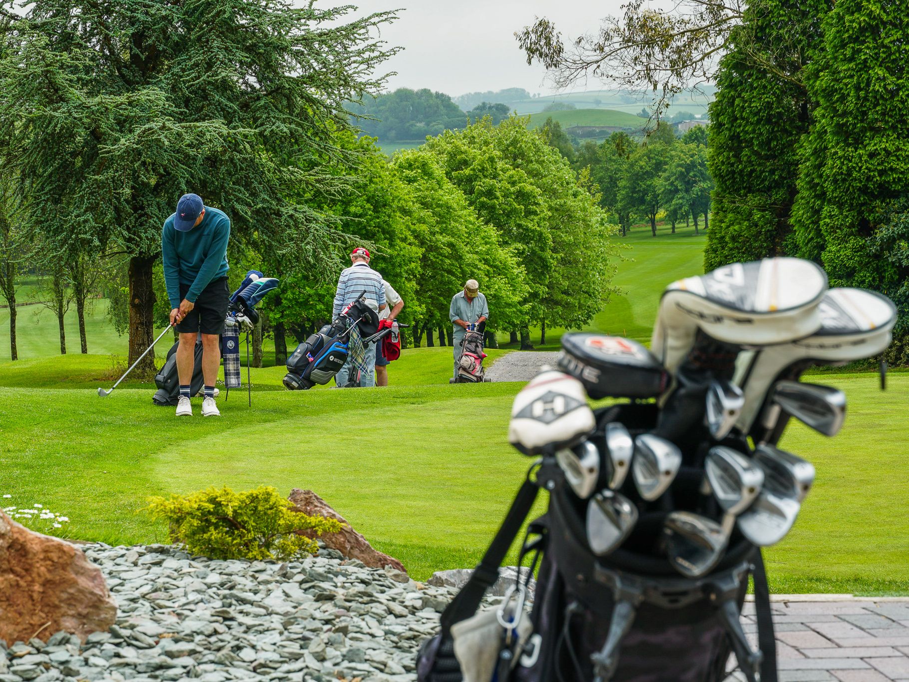 Golfers preparing to play on a green golf course with golf bags and clubs in the foreground. Trees and rolling hills are visible in the background.