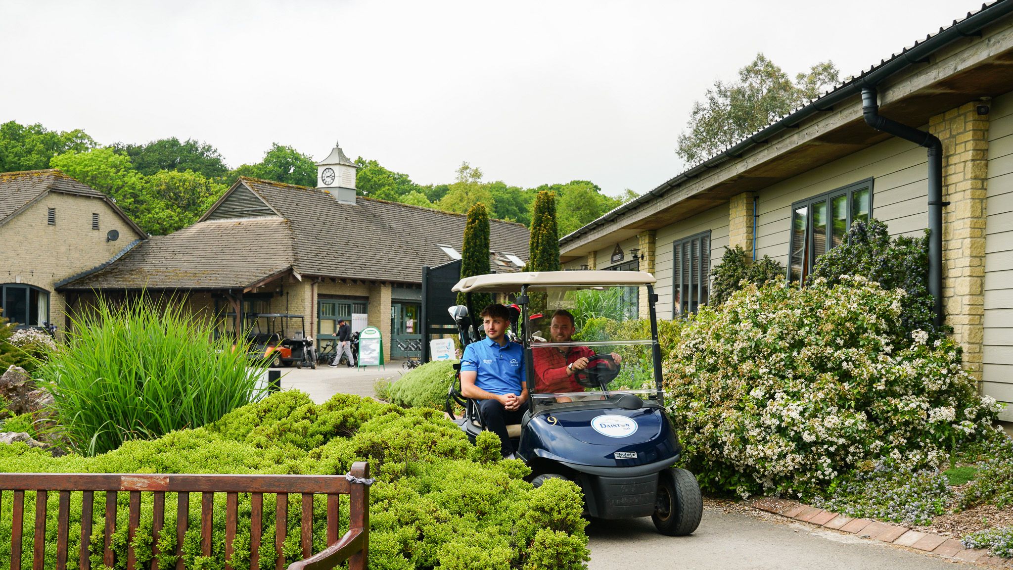 Two people sitting in a golf cart outside a clubhouse surrounded by greenery, with a wooden bench and blooming bushes in the foreground.