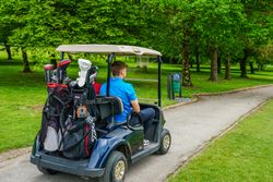 Man riding a golf cart with golf bags on a paved path through a green, tree-filled golf course.