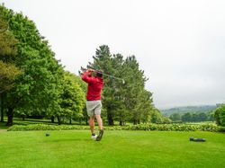Person in a red jacket and shorts swinging a golf club on a green golf course surrounded by trees and hills under a cloudy sky.