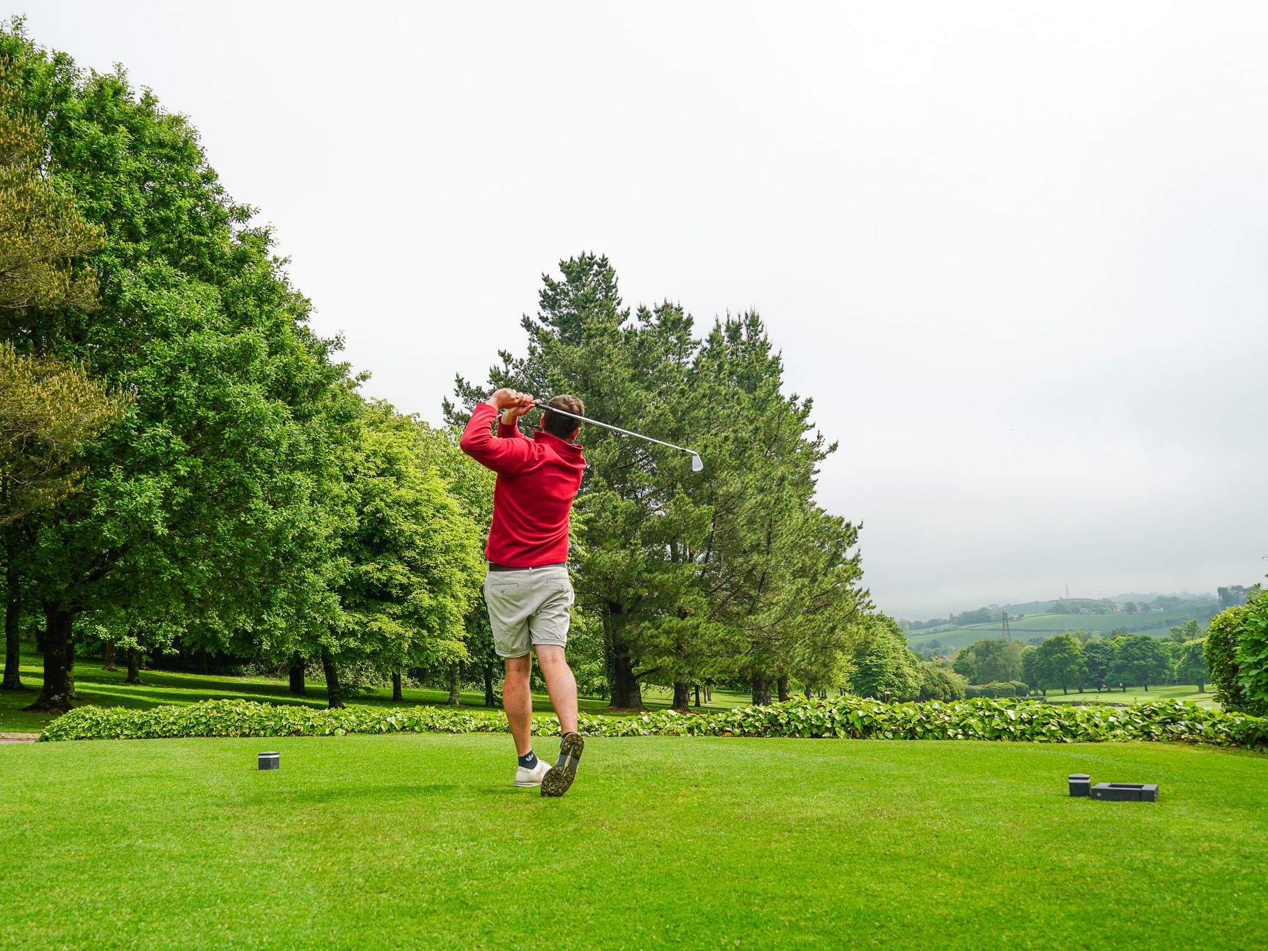 Person in a red jacket and shorts swinging a golf club on a green golf course surrounded by trees and hills under a cloudy sky.