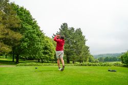 Person in a red jacket and shorts swinging a golf club on a green golf course surrounded by trees and hills under a cloudy sky.