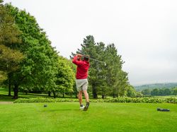 Person in a red jacket and shorts swinging a golf club on a green golf course surrounded by trees and hills under a cloudy sky.