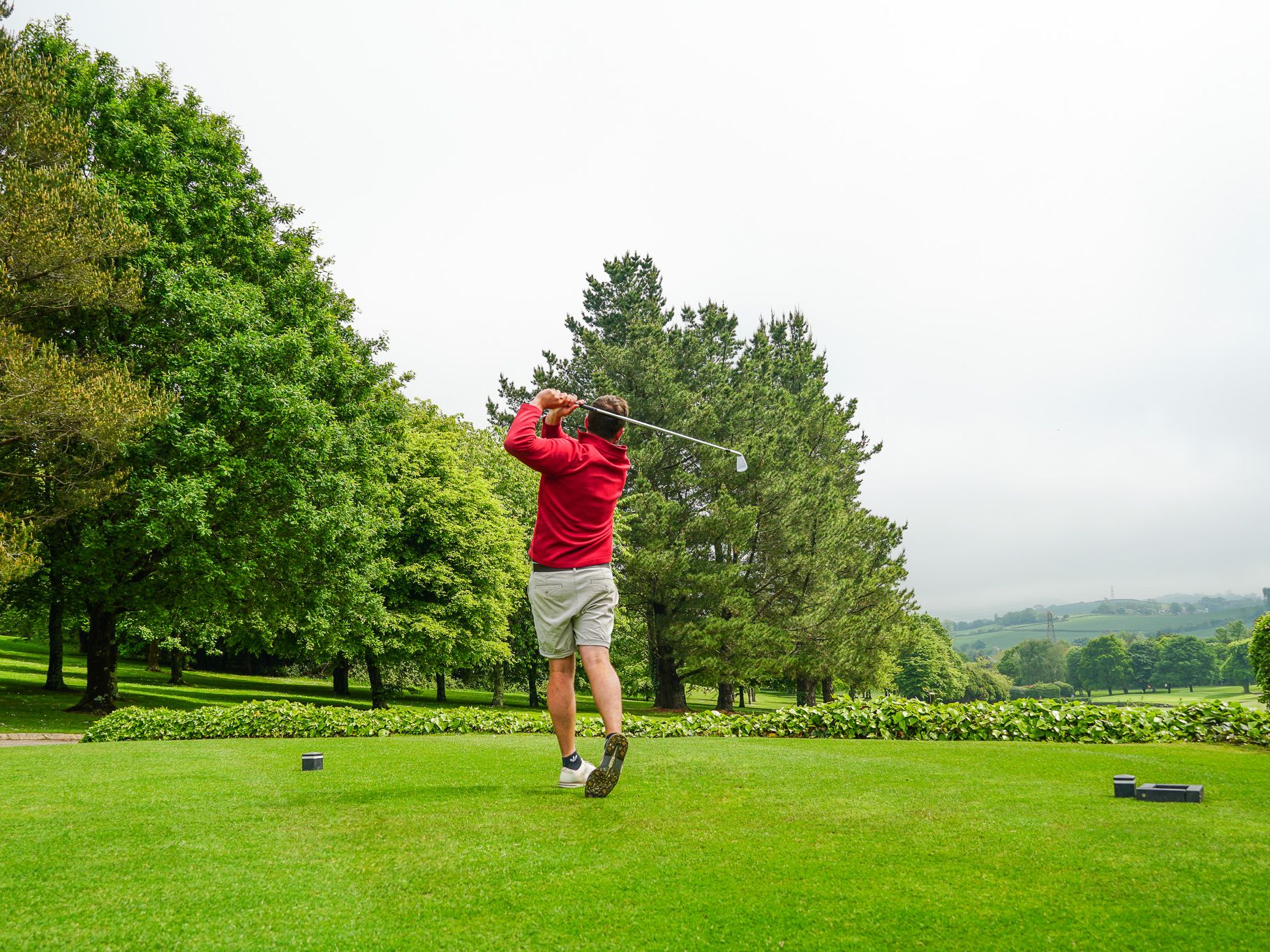 Person in a red jacket and shorts swinging a golf club on a green golf course surrounded by trees and hills under a cloudy sky.