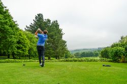 Person playing golf, swinging a club on a lush green golf course with trees and hills in the background.