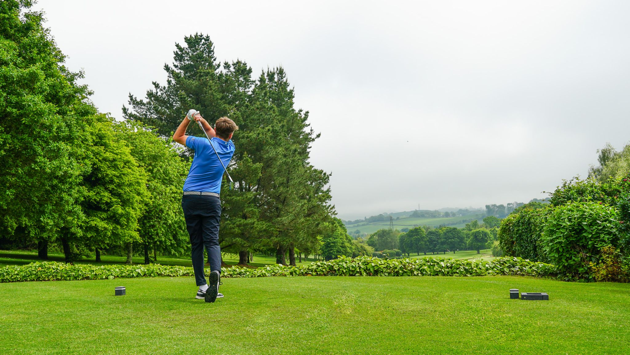 Person playing golf, swinging a club on a lush green golf course with trees and hills in the background.